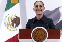 President Sheinbaum smiles from behind a podium next to a Mexican flag