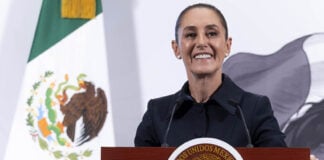 President Sheinbaum smiles from behind a podium next to a Mexican flag