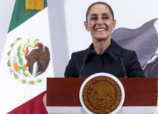 President Sheinbaum smiles from behind a podium next to a Mexican flag