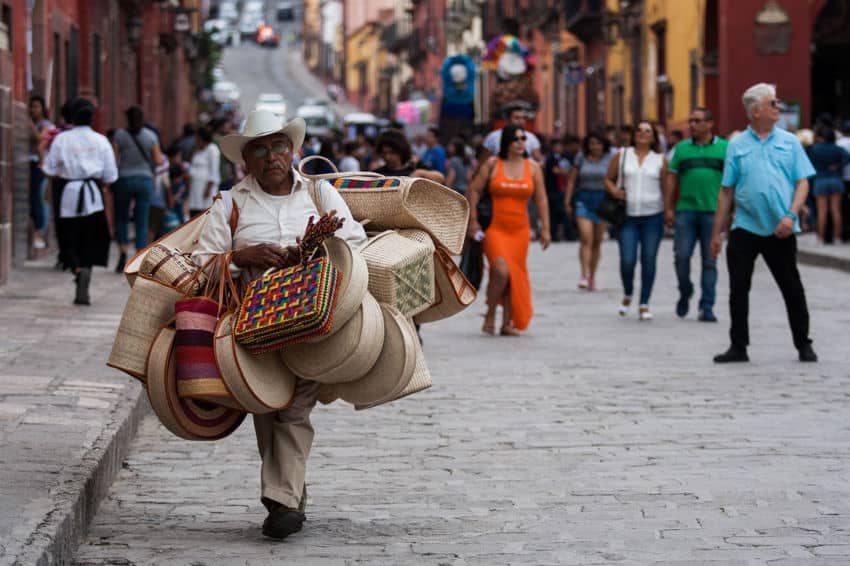 An artisan street vendor, wearing a traditional straw cowboy hat, walks along a bustling cobblestone street in San Miguel de Allende. He carries an load of handmade woven palm baskets, hats, and colorful bags for sale on his shoulders.