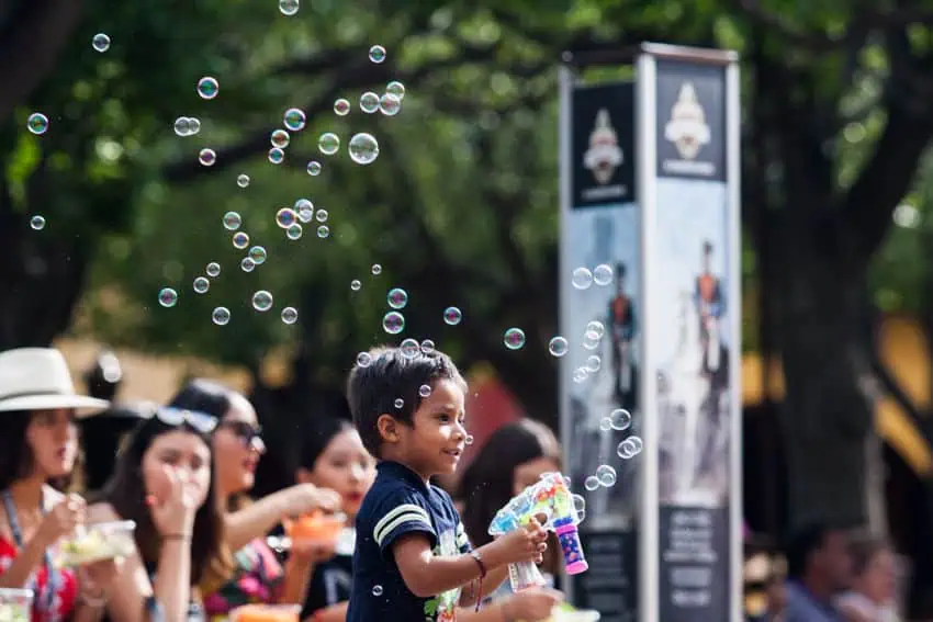 A young boy smiles as he plays with a bubble gun in a sunny public plaza in San Miguel de Allende. He is surrounded by adults sitting on park benches in the background.