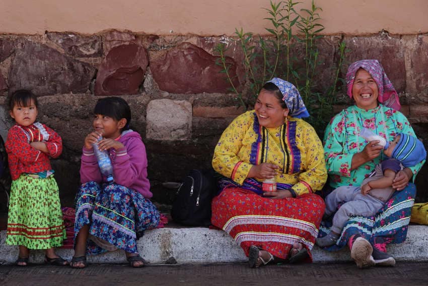 A group of Rarámuri (Tarahumara) women and children resting together against a rustic stone wall. They are dressed in traditional vibrant clothing; the two women are laughing, while one holds a baby drinking from a bottle, and two young girls sit beside them.