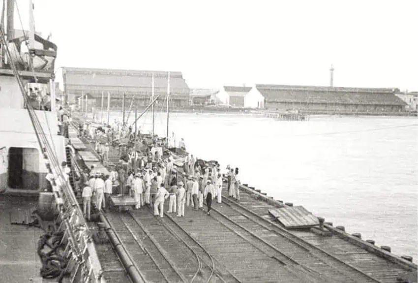 Black and white photo of a crowded dockside scenewith men dressed in white clothing and hats, standing on a wooden pier next to a large ship in Mexico. Some are pushing flatbed carts on railroad tracks that run along the pier. In the background, there is a large body of water and several industrial-looking buildings.