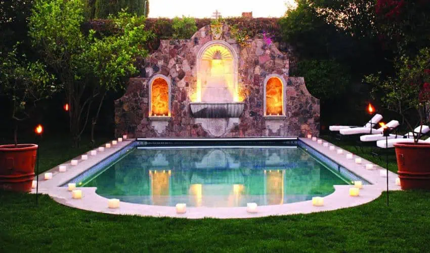 A candlelit swimming pool at a luxury boutique hotel in San Miguel de Allende, illuminated at dusk with torches around the pool. A colonial-style stone wall with a softly lit fountain is in the background.