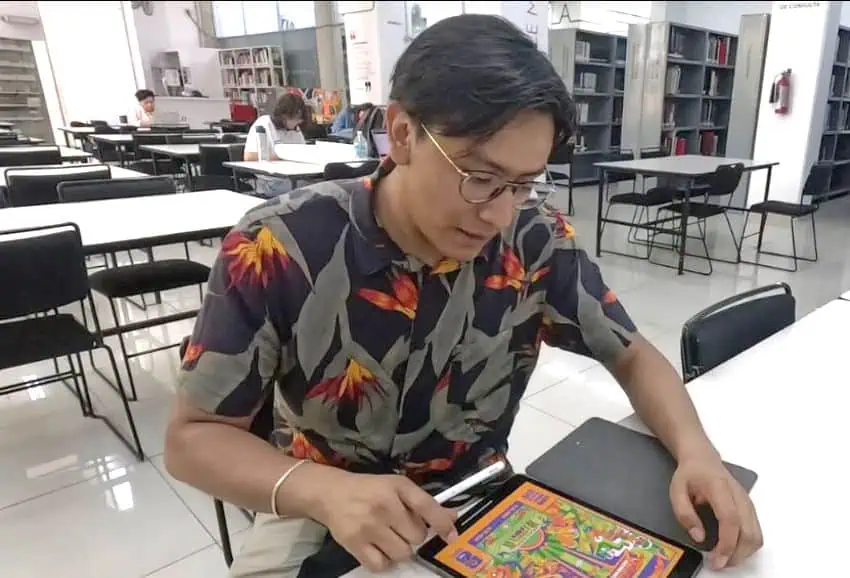 Mexican designer Mario Cortés, wearing glasses and a patterned shirt, sits at a white table inside a library. He is looking down at a tablet, which displays one of his colorful and intricate digital illustrations, while holding a stylus.