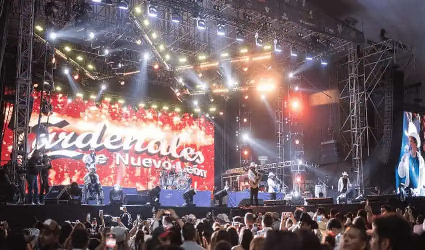 A live concert in Mexico with a large band on a stage illuminated by bright lights. The band, Los Cardenales de Nuevo León, is playing to a large crowd of people holding up their phones to record the performance.