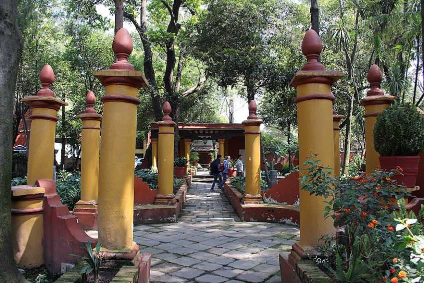 A stone flagstone pathway at the Fonoteca Nacional gardens in Mexico City. The path is flanked by rows of thick, mustard-yellow pillars with decorative reddish-brown tops, all set within a lush, green wooded area.