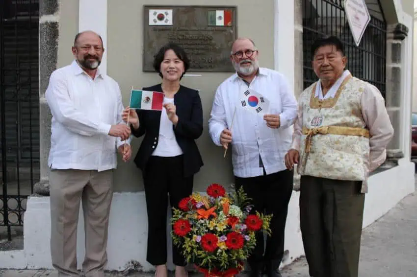 A color photo of four government officials of Korea and Mexico osing outdoors in Merida, Mexico, in front of a bronze plaque. The person on the far left and the second from the right are men holding small Mexican and South Korean flags. They are dressed in white shirts and light-colored trousers. The woman in the center is holding a Mexican flag and is dressed in a dark blazer and white blouse. The man on the far right is wearing traditional Korean clothing, including a hanbok-style vest. A wreath of flowers sits on the ground in front of the group.