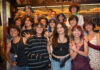 A diverse group of smiling Mexican teenagers, with various hairstyles and clothing styles, pose for a group photo in a dimly lit restaurant or bar with rows of candles in the background.