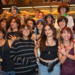 A diverse group of smiling Mexican teenagers, with various hairstyles and clothing styles, pose for a group photo in a dimly lit restaurant or bar with rows of candles in the background.