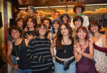 A diverse group of smiling Mexican teenagers, with various hairstyles and clothing styles, pose for a group photo in a dimly lit restaurant or bar with rows of candles in the background.