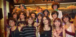 A diverse group of smiling Mexican teenagers, with various hairstyles and clothing styles, pose for a group photo in a dimly lit restaurant or bar with rows of candles in the background.