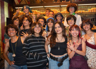 A diverse group of smiling Mexican teenagers, with various hairstyles and clothing styles, pose for a group photo in a dimly lit restaurant or bar with rows of candles in the background.