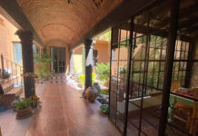 A colonial-style covered corridor with terracotta tile floors, arched brick ceilings, and large windows overlooking a courtyard garden in San Miguel de Allende.