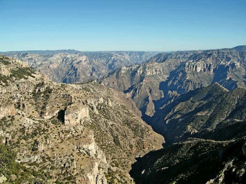 A panoramic view of Mexico's Copper Canyon in Chihuahua. The rugged canyon system stretches out under a clear blue sky, showing deep gorges and rocky, scrub-covered mesas.