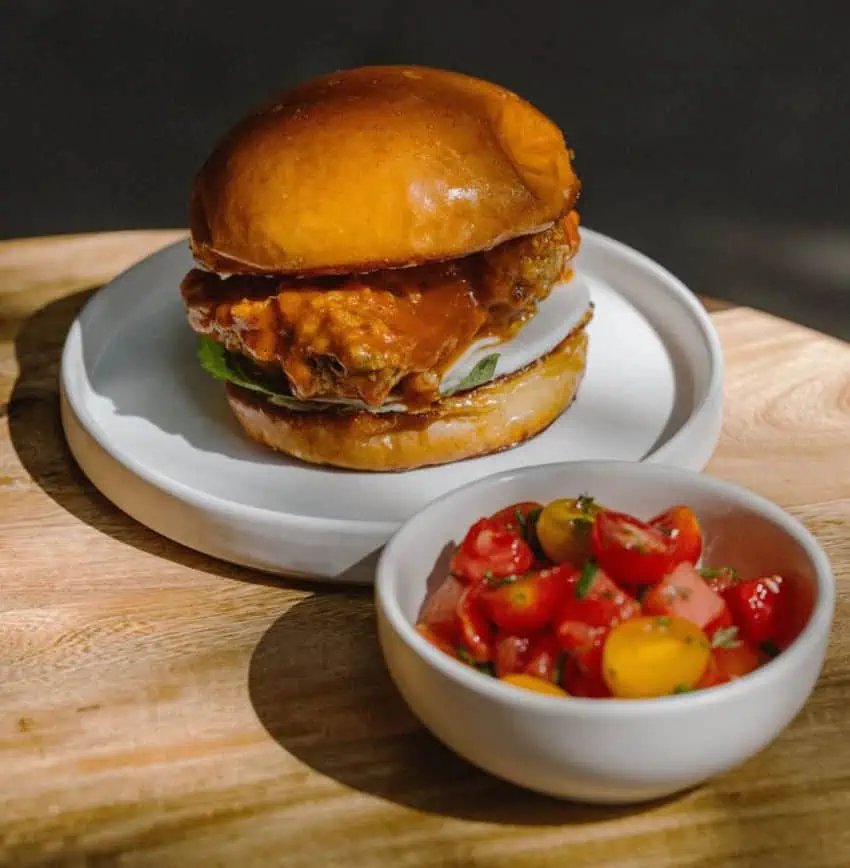 A juicy-looking chicken sandwich with a golden-brown bun sits on a white plate. Next to it, a small white bowl holds a vibrant salad of red and yellow cherry tomatoes. Both dishes are placed on a light wooden cutting board.