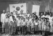 Black and white photo of a group of Mexican Koreans and Maya people posing together in front of a wall bearing two flags — one Korean and another a banner reminiscent of the Mexican flag. The group includes several adults and many children. The adults are standing in the back row while the children are in front. The subjects are dressed in simple, everyday clothing.