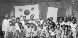 Black and white photo of a group of Mexican Koreans and Maya people posing together in front of a wall bearing two flags — one Korean and another a banner reminiscent of the Mexican flag. The group includes several adults and many children. The adults are standing in the back row while the children are in front. The subjects are dressed in simple, everyday clothing.