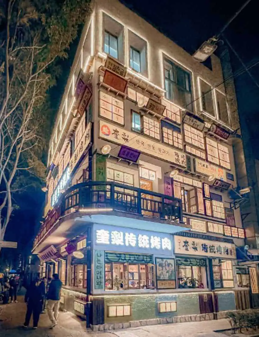 A multi-story building is shown at night. It is brightly lit with numerous windows and signs on its exterior. People are walking on the sidewalk in front of the building. The trees on the left side are bare, suggesting it is winter or a cooler season.