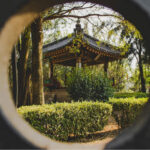 A circular framing shows a Japanese-style gazebo with a dark tiled roof and ornate trim, nestled among trees and manicured hedges in a park.