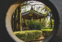 A circular framing shows a Japanese-style gazebo with a dark tiled roof and ornate trim, nestled among trees and manicured hedges in a park.