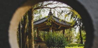 A circular framing shows a Japanese-style gazebo with a dark tiled roof and ornate trim, nestled among trees and manicured hedges in a park.