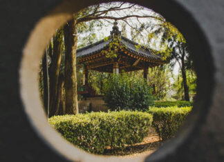 A circular framing shows a Japanese-style gazebo with a dark tiled roof and ornate trim, nestled among trees and manicured hedges in a park.