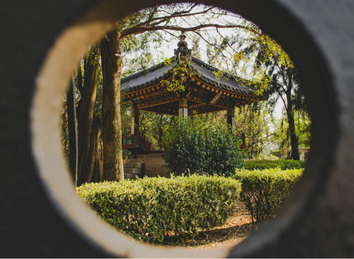 A circular framing shows a Japanese-style gazebo with a dark tiled roof and ornate trim, nestled among trees and manicured hedges in a park.