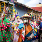 Costumed dancers in vibrant masks and traditional clothing parade through the streets during a Mexican festival in Xico, Veracruz, with colorful banners and colonial buildings in the background.