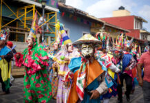 Costumed dancers in vibrant masks and traditional clothing parade through the streets during a Mexican festival in Xico, Veracruz, with colorful banners and colonial buildings in the background.