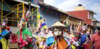 Costumed dancers in vibrant masks and traditional clothing parade through the streets during a Mexican festival in Xico, Veracruz, with colorful banners and colonial buildings in the background.