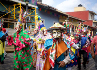 Costumed dancers in vibrant masks and traditional clothing parade through the streets during a Mexican festival in Xico, Veracruz, with colorful banners and colonial buildings in the background.
