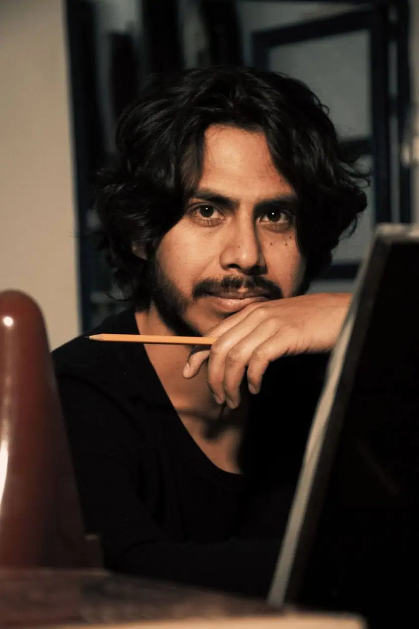 A man with dark wavy hair and a goatee looks thoughtfully at the camera. He is resting his chin on his hand and holding a yellow pencil, seated behind an easel in a warmly lit, artistic setting.