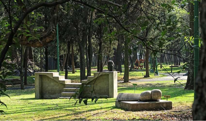 A tranquil park scene with various modern art sculptures scattered across a green lawn, surrounded by tall trees and dappled sunlight.
