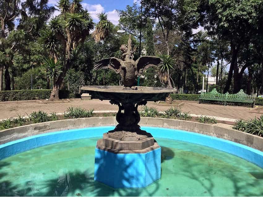 An ornate fountain at Parque Lira in Mexico City. A stone sculpture of a child riding a swan sits on a bright blue base within a turquoise-painted pool, surrounded by park benches and trees.