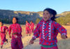 A group of young Rarámuri girls participate in a traditional running game in a rural, mountainous landscape under a clear sky. They are wearing brightly colored pink dresses with blue floral patterns and white zigzag trim, and some are holding sticks. The girl in the foreground looks directly at the viewer