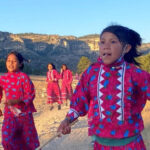 A group of young Rarámuri girls participate in a traditional running game in a rural, mountainous landscape under a clear sky. They are wearing brightly colored pink dresses with blue floral patterns and white zigzag trim, and some are holding sticks. The girl in the foreground looks directly at the viewer