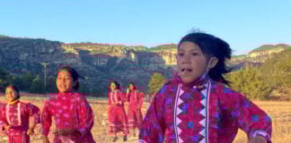 A group of young Rarámuri girls participate in a traditional running game in a rural, mountainous landscape under a clear sky. They are wearing brightly colored pink dresses with blue floral patterns and white zigzag trim, and some are holding sticks. The girl in the foreground looks directly at the viewer