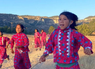 A group of young Rarámuri girls participate in a traditional running game in a rural, mountainous landscape under a clear sky. They are wearing brightly colored pink dresses with blue floral patterns and white zigzag trim, and some are holding sticks. The girl in the foreground looks directly at the viewer