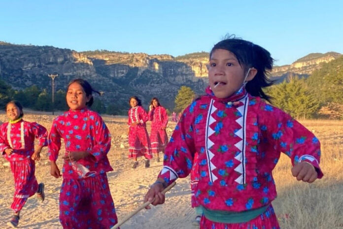 A group of young Rarámuri girls participate in a traditional running game in a rural, mountainous landscape under a clear sky. They are wearing brightly colored pink dresses with blue floral patterns and white zigzag trim, and some are holding sticks. The girl in the foreground looks directly at the viewer