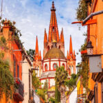 A vibrant photo of the pink neo-Gothic spires of the Parroquia de San Miguel Arcángel in San Miguel de Allende, Mexico, viewed from a city street.