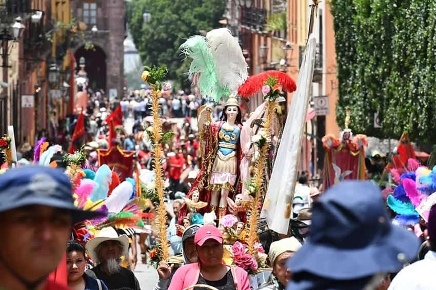 A street-level view of a crowded religious procession for the feast day of San Miguel Arcángel in San Miguel de Allende, Mexico. A statue of the patron saint is carried on a platform, surrounded by participants in the parade and traditional, colorful feather decorations.