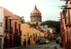 A warm, late-afternoon street scene in San Miguel de Allende, Mexico. Colorful colonial buildings in shades of pink and yellow line a cobblestone street where people are walking. The iconic dome of the Las Monjas church is visible in the background against a pale sky.