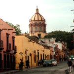 A warm, late-afternoon street scene in San Miguel de Allende, Mexico. Colorful colonial buildings in shades of pink and yellow line a cobblestone street where people are walking. The iconic dome of the Las Monjas church is visible in the background against a pale sky.