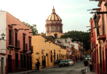 A warm, late-afternoon street scene in San Miguel de Allende, Mexico. Colorful colonial buildings in shades of pink and yellow line a cobblestone street where people are walking. The iconic dome of the Las Monjas church is visible in the background against a pale sky.