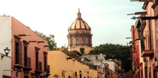 A warm, late-afternoon street scene in San Miguel de Allende, Mexico. Colorful colonial buildings in shades of pink and yellow line a cobblestone street where people are walking. The iconic dome of the Las Monjas church is visible in the background against a pale sky.