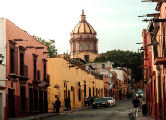 A warm, late-afternoon street scene in San Miguel de Allende, Mexico. Colorful colonial buildings in shades of pink and yellow line a cobblestone street where people are walking. The iconic dome of the Las Monjas church is visible in the background against a pale sky.