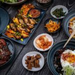 A variety of Korean dishes are arranged on a dark, textured table. The spread includes grilled meats, a large bibimbap bowl with a fried egg, various small side dishes (banchan) like kimchi and pickled vegetables, and a savory pancake. The plates are dark blue and black, contrasting with the vibrant colors of the food.