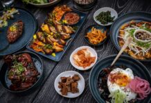 A variety of Korean dishes are arranged on a dark, textured table. The spread includes grilled meats, a large bibimbap bowl with a fried egg, various small side dishes (banchan) like kimchi and pickled vegetables, and a savory pancake. The plates are dark blue and black, contrasting with the vibrant colors of the food.