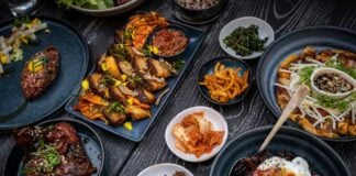 A variety of Korean dishes are arranged on a dark, textured table. The spread includes grilled meats, a large bibimbap bowl with a fried egg, various small side dishes (banchan) like kimchi and pickled vegetables, and a savory pancake. The plates are dark blue and black, contrasting with the vibrant colors of the food.